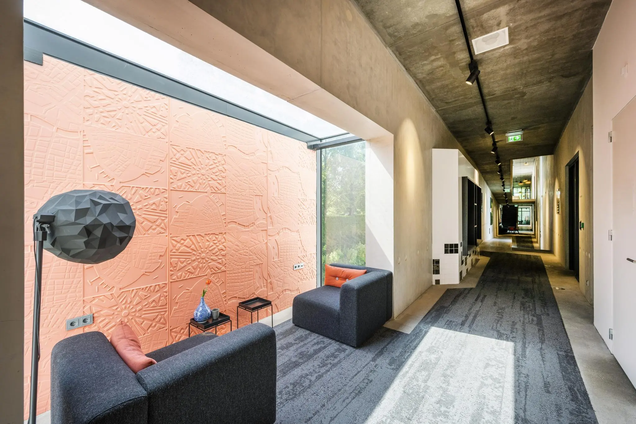 Moderne gang met betonnen plafond, zitgedeelte bij een groot raam en oranje wanddecoratie met reliëfpatroon aan de Fred. Roeskestraat.